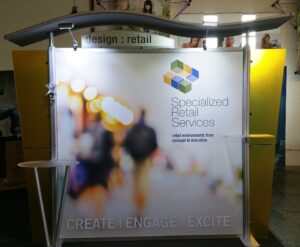 A trade show booth for "Specialized Retail Services" features a prominently displayed logo and tagline. The bright display showcases a blurred background of a retail environment with "CREATE | ENGAGE | EXCITE" written at the bottom.