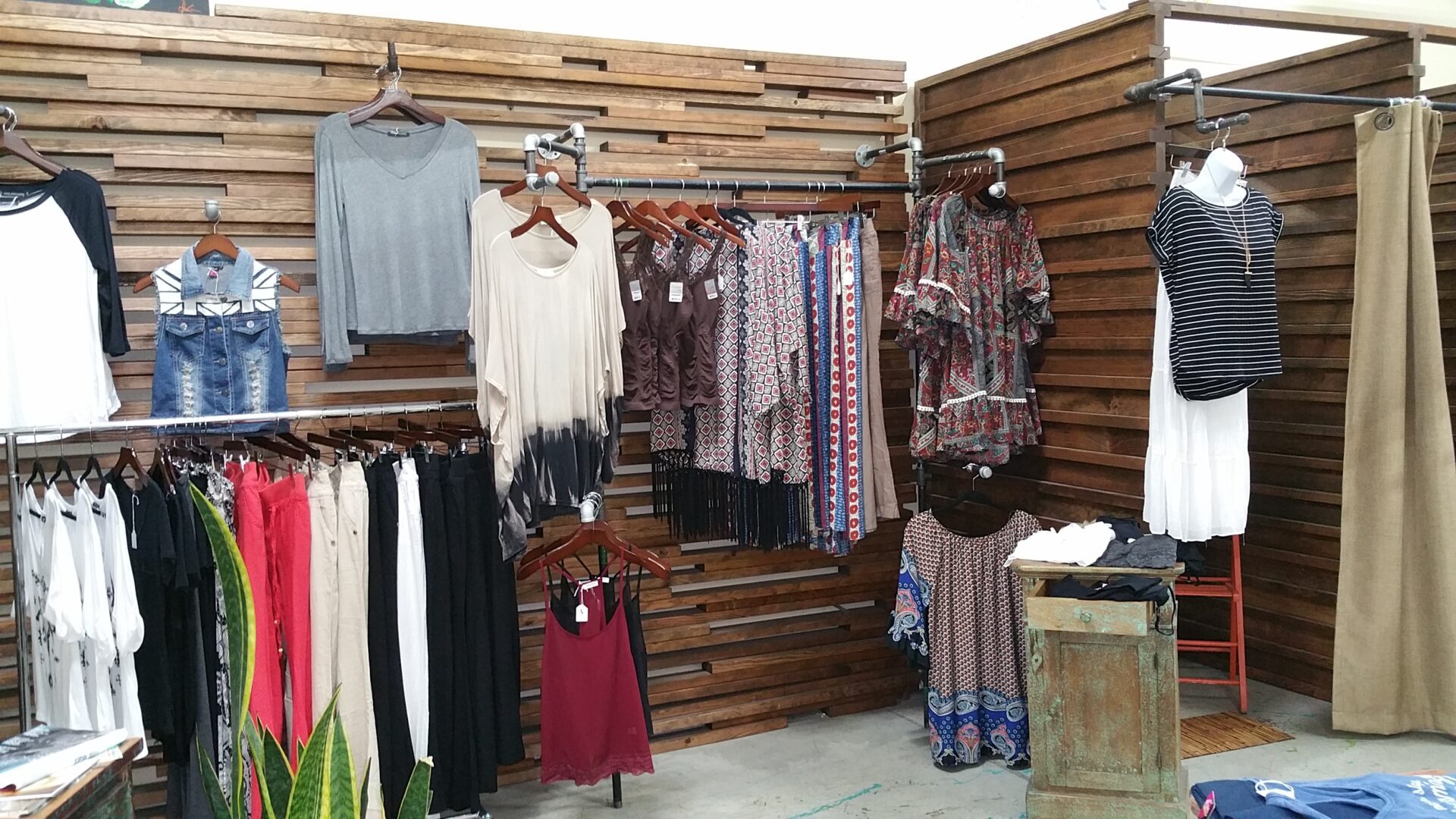 A boutique clothing store displays various garments on wooden racks and hangers. Visible items include patterned skirts, blouses, dresses, and pants in a mix of casual and eclectic styles. Potted plants and a vintage dresser add decorative touches to the space.