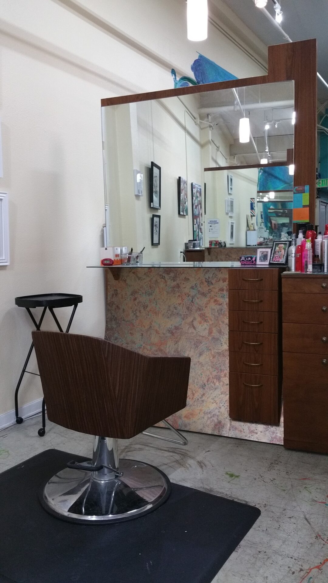 A salon station with a large mirror, a wooden chair with a metal base, and a counter holding various hair products and picture frames. The station includes a set of drawers and a side cart. The background shows an art-adorned wall and other salon stations.