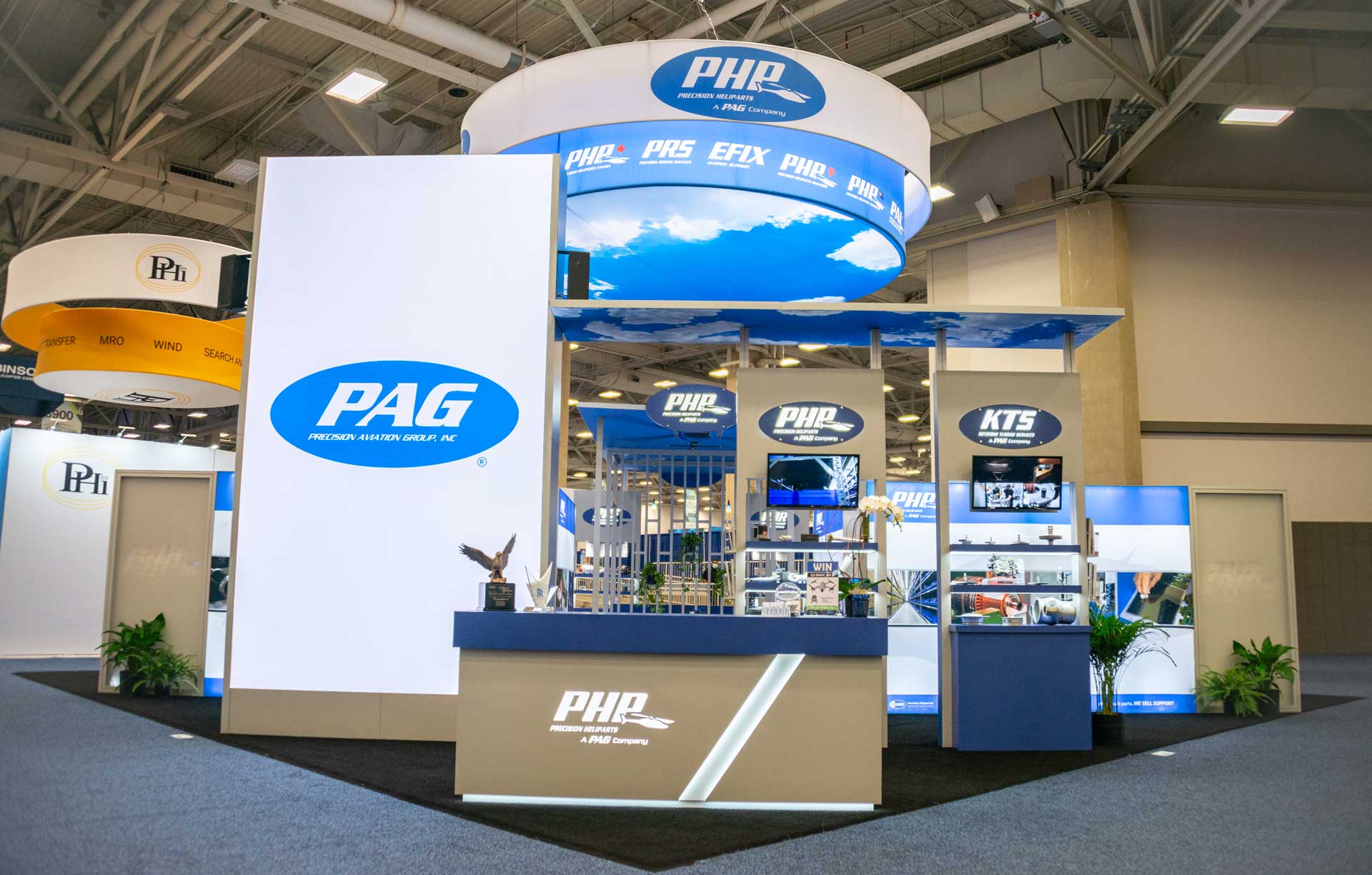 An exhibition booth with a primary blue and white color scheme featuring large signs with "PAG" and "PHP" logos. The booth has a reception desk, multiple display screens, and promotional materials. The surrounding area has other booths and an industrial ceiling.