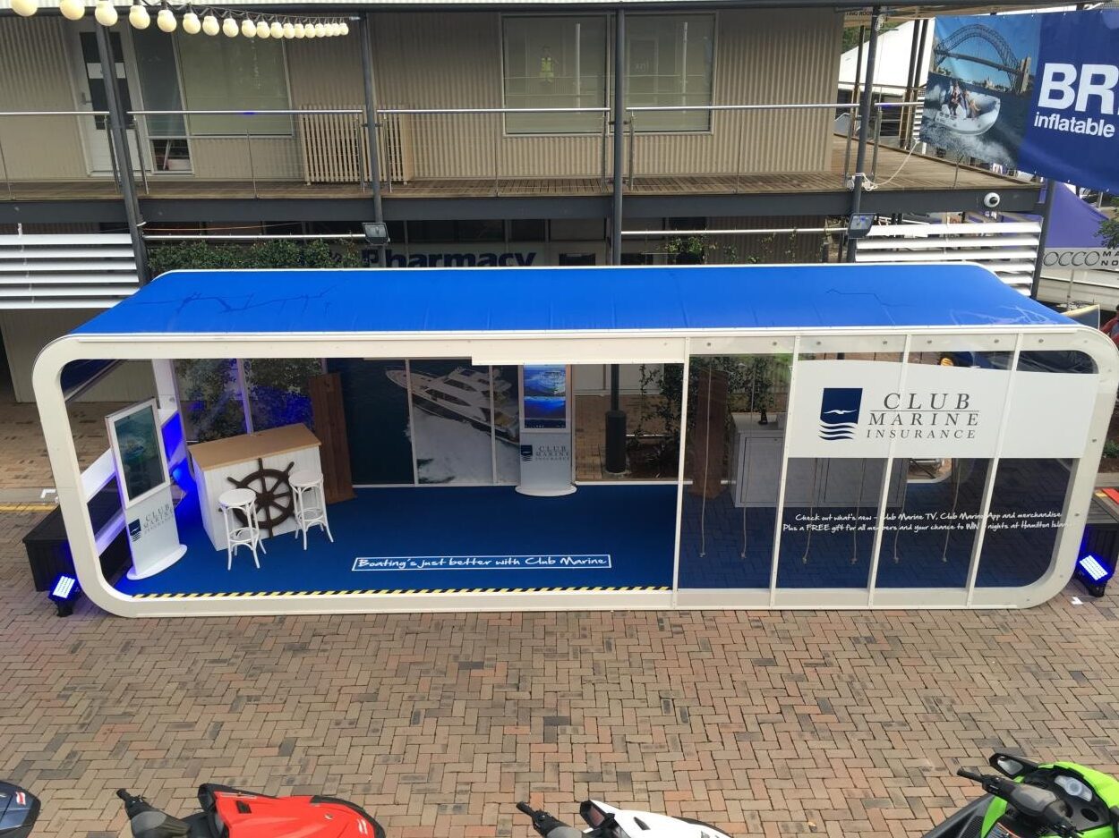 A pop-up container booth for Club Marine Insurance is seen, featuring a blue and white color scheme. Inside, there is a table with chairs and promotional materials. The booth is set outdoors on a brick pavement with a building and pharmacy sign in the background.