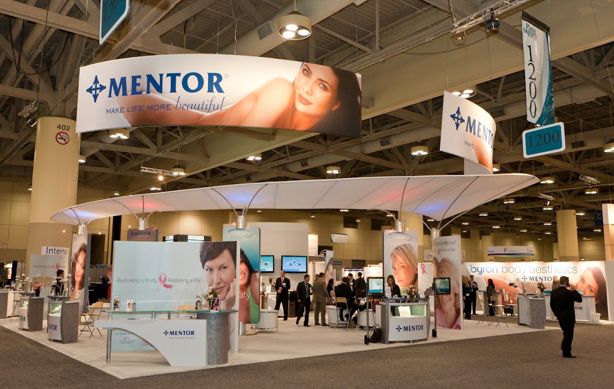 A well-lit exhibition booth at a convention center is branded with the name “Mentor” and the tagline “Make Life More Beautiful.” The booth features multiple display counters, banners with photos, and several people interacting around the area.