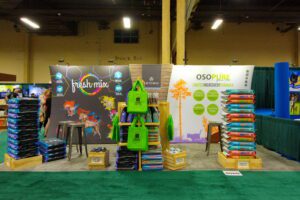 A trade show booth features a colorful display with pet food products. Three standing banners showcase branding for "Fresh Mix" and "Osopure Limited Ingredient Formula." Green reusable bags and stacked pet food bags are also prominently displayed.