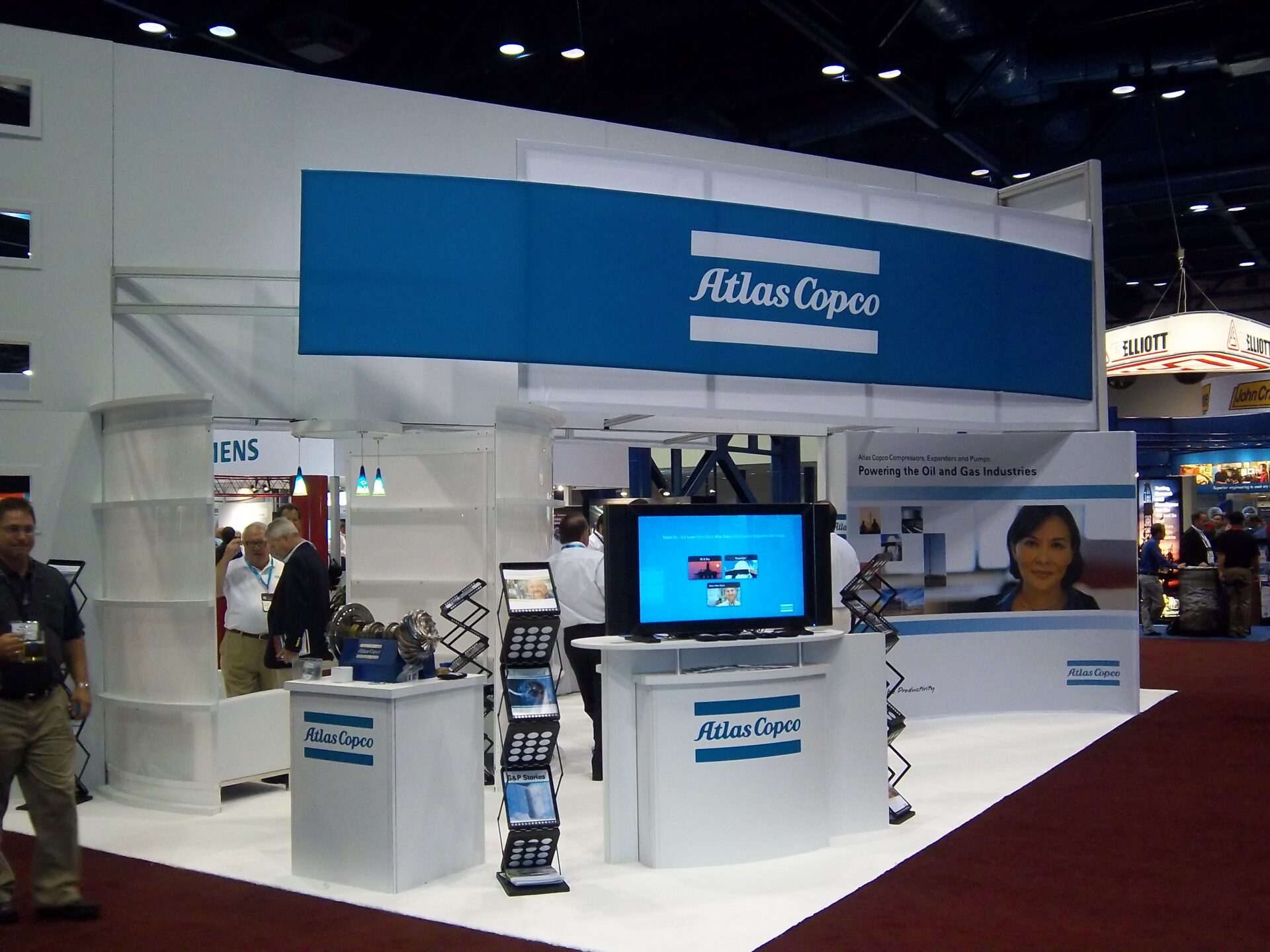 A trade show booth features a prominent blue and white "Atlas Copco" sign. The booth includes displays with promotional materials, a TV screen, shelves, and a counter. Several people are present, engaging with the booth setup or walking nearby.