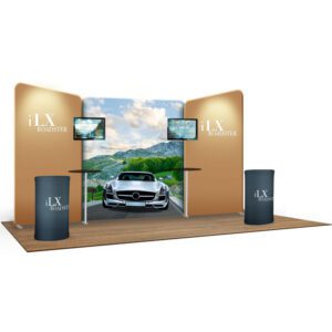 A trade show booth displays "iLX Roadster" with a backdrop featuring a white sports car driving on a scenic mountain road. Two flat screens are mounted on either side of the backdrop. The booth includes two round, branded counters and a wooden floor platform.