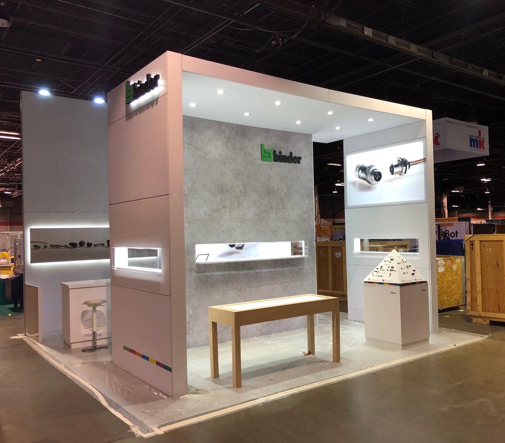 Exhibition booth with a modern design featuring a tall, white structure with the name 'Binder'. The booth has display areas showcasing various products, a small wooden table, and a white stool. The surrounding area is dimly lit with other booths visible in the background.