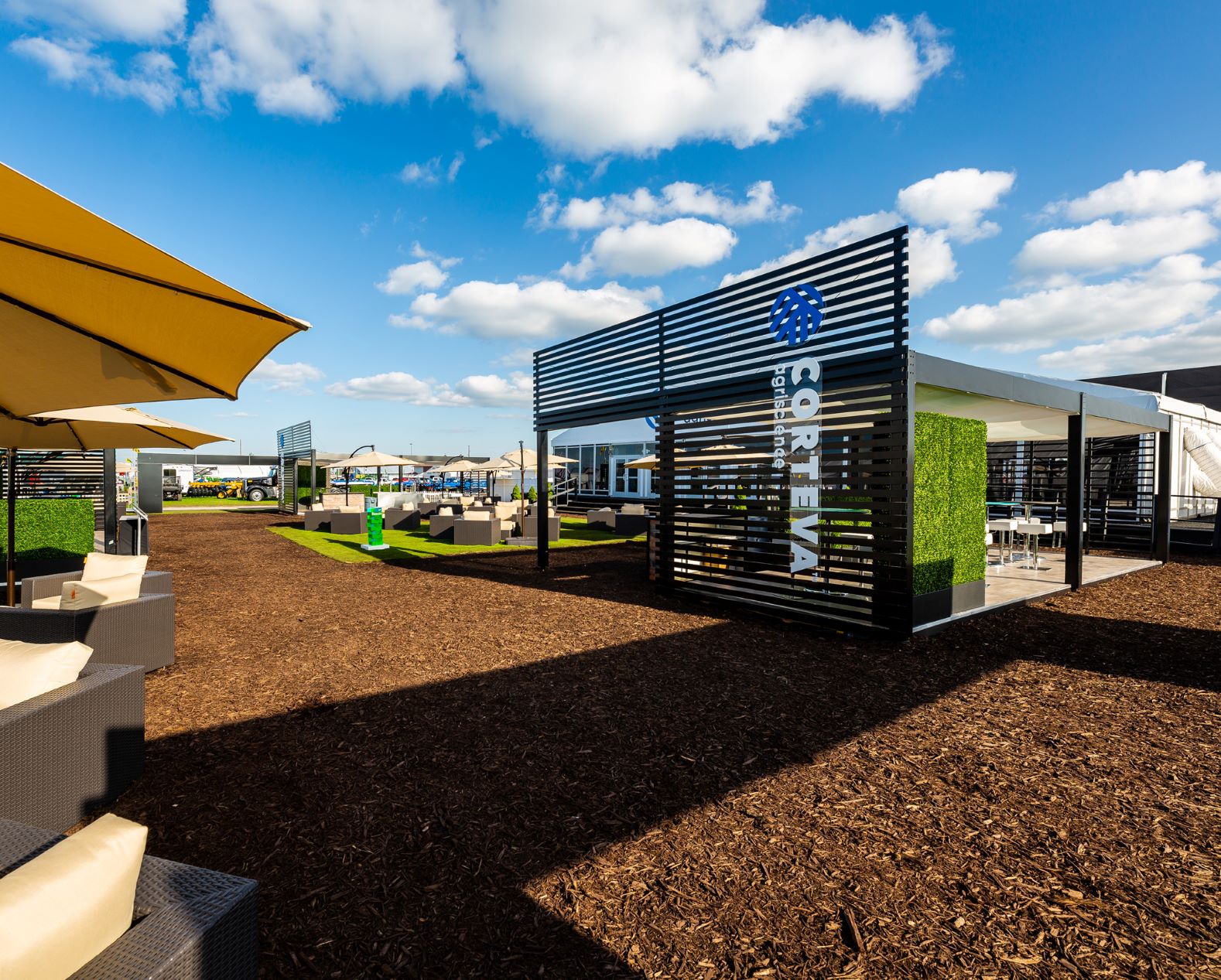 An outdoor seating area with modern, partially enclosed canopies at the Syngenta SCREWVAL facility. The area features chairs, tables, and green artificial turf, under a bright, sunny sky with scattered clouds. Large umbrellas provide additional shade.