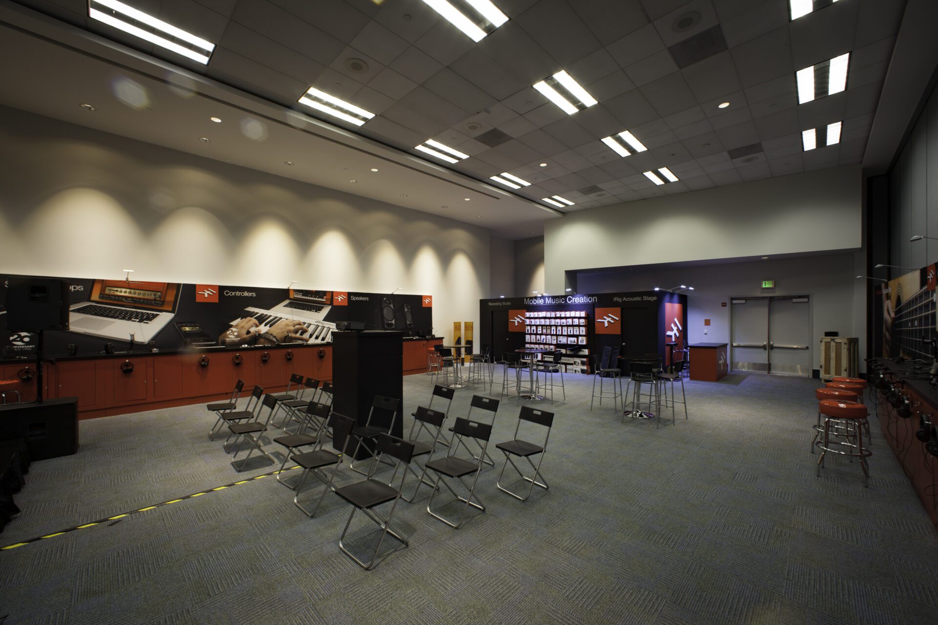 A conference room with rows of chairs facing a presentation area. The room has a high ceiling with bright lighting. Various equipment is displayed on stands along the walls. There is a door in the right corner and the floor is carpeted in blue.