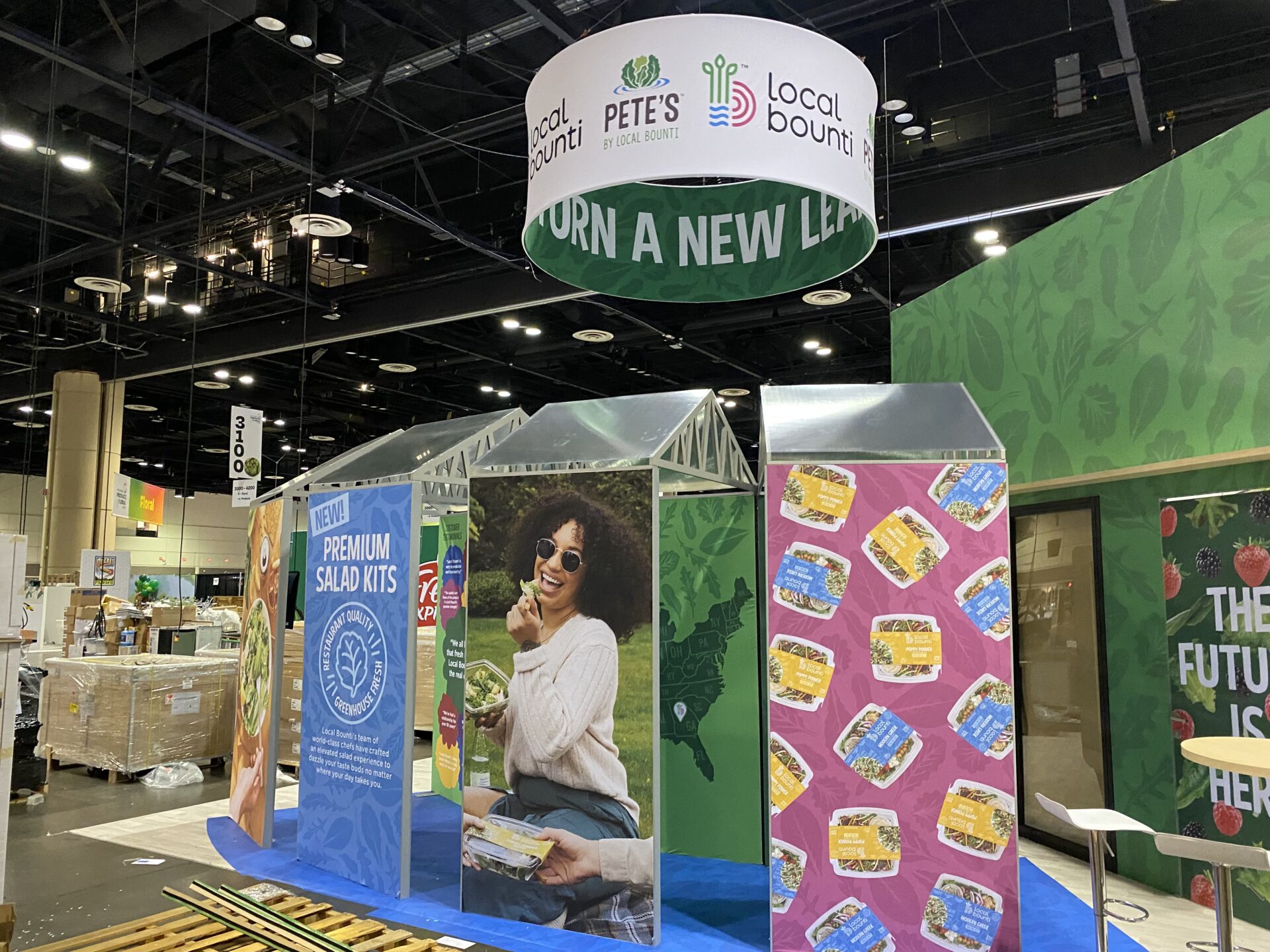 A booth display at a trade show features the Local Bounti and Pete's brands. It includes images of salad kits, colorful posters, and a large banner overhead. A sign on the left reads "NEW! Premium Salad Kits." The setup appears bright and inviting with green and blue colors.