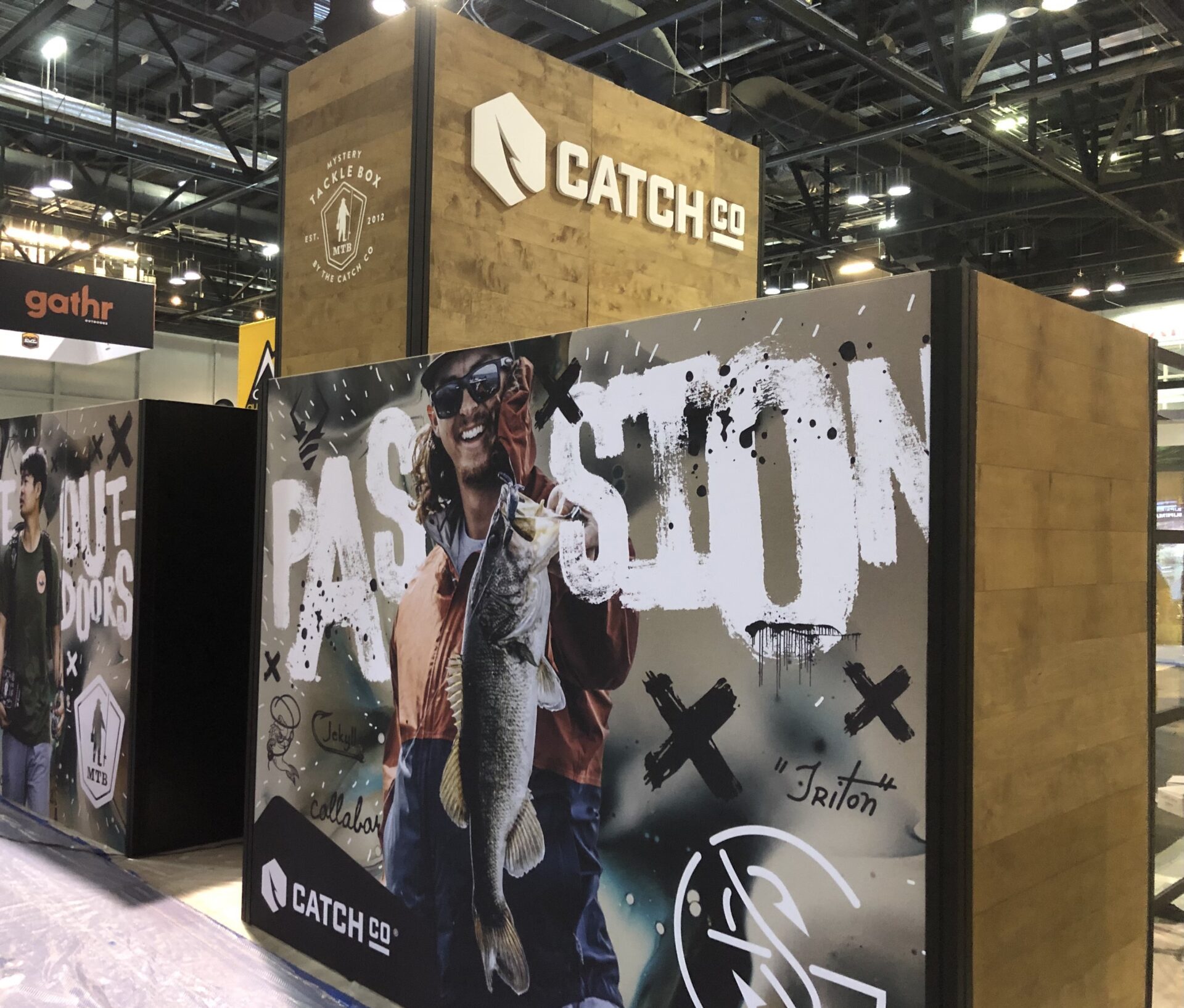 A display booth at a trade show featuring Catch Co. branding. The booth has a large wooden structure with the company logo. A banner shows a person holding a fish with the word "PASSION" prominently displayed in white text. Other booths and signs are visible in the background.