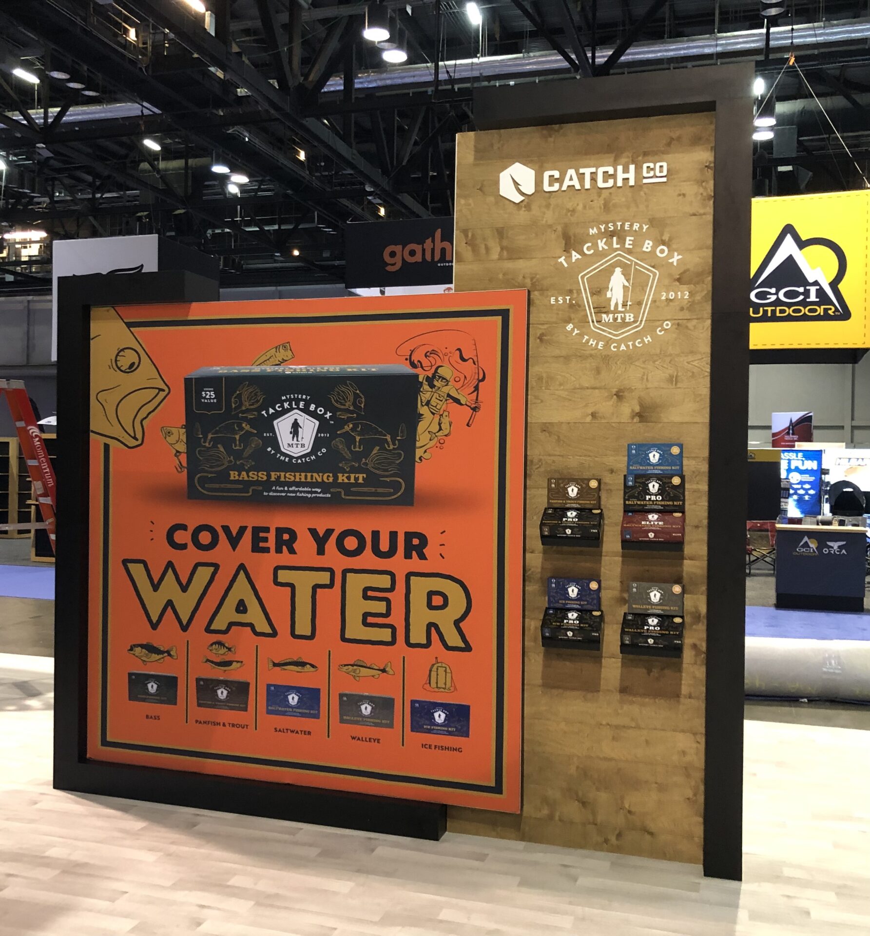 A trade show booth for Catch Co. features a display of their Mystery Tackle Box products. The backdrop includes imagery of fish and reads "Cover Your Water." Various fishing kits are showcased on shelves against a wood panel background.