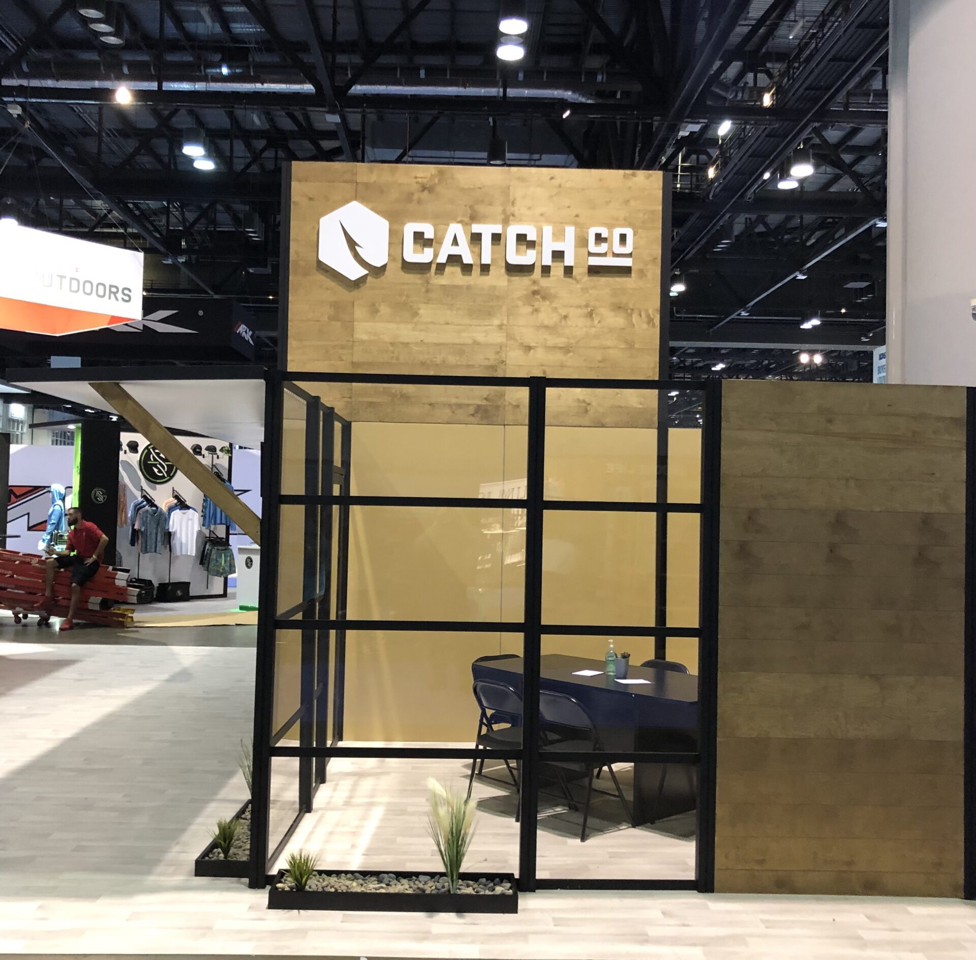 Indoor trade show booth with a wood panel background displaying the text "CATCH CO" under a geometric logo. The booth has a glass-walled area with a round table and chairs. Green plants are arranged at the base of the glass panels.