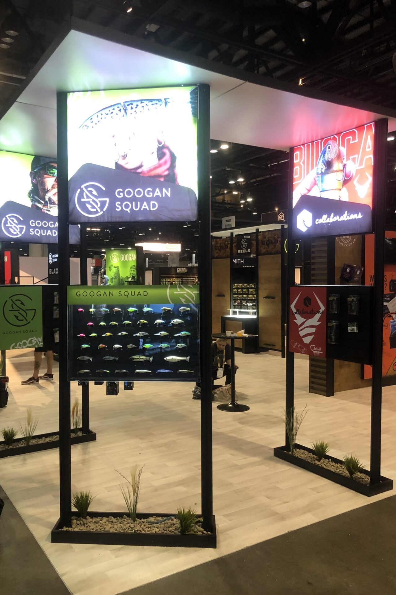 A well-lit trade show booth features the Googan Squad, showcasing their fishing gear. The booth has illuminated signs, display racks with products, and informational panels. The setup includes interactive displays and promotional graphics under a high ceiling.