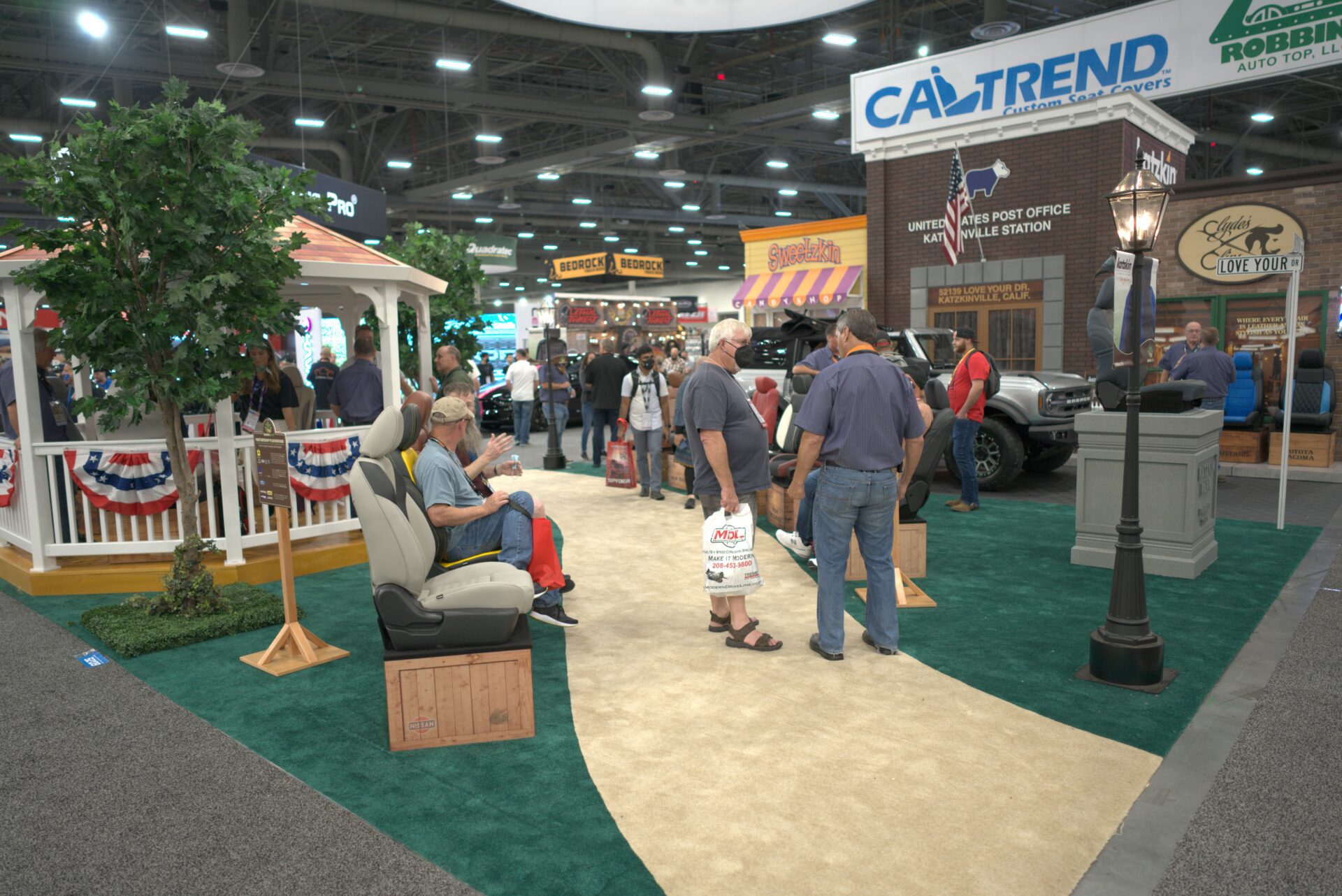 A trade show or expo featuring multiple booths and a crowd of attendees. Some people are walking and others are gathered near booths showcasing various products, including automotive accessories. Decorative elements like a gazebo, streetlamp, and a tree are present.