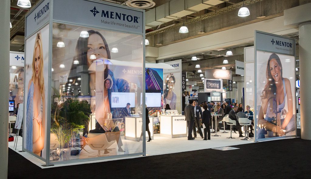 A large, well-lit exhibition booth for Mentor featuring smiling models in the background. People are seen engaging in discussions around tables within the booth. The banners display the slogan "Make life more beautiful." The surrounding area is bustling with attendees.