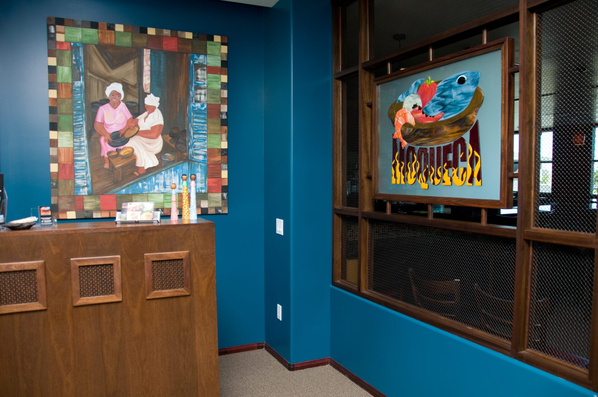 A brightly lit room with blue walls features two colorful paintings. One painting depicts two people, and the other shows a heart surrounded by birds. There is a wooden reception desk with a mosaic pattern, and part of a seating area is visible through a window.