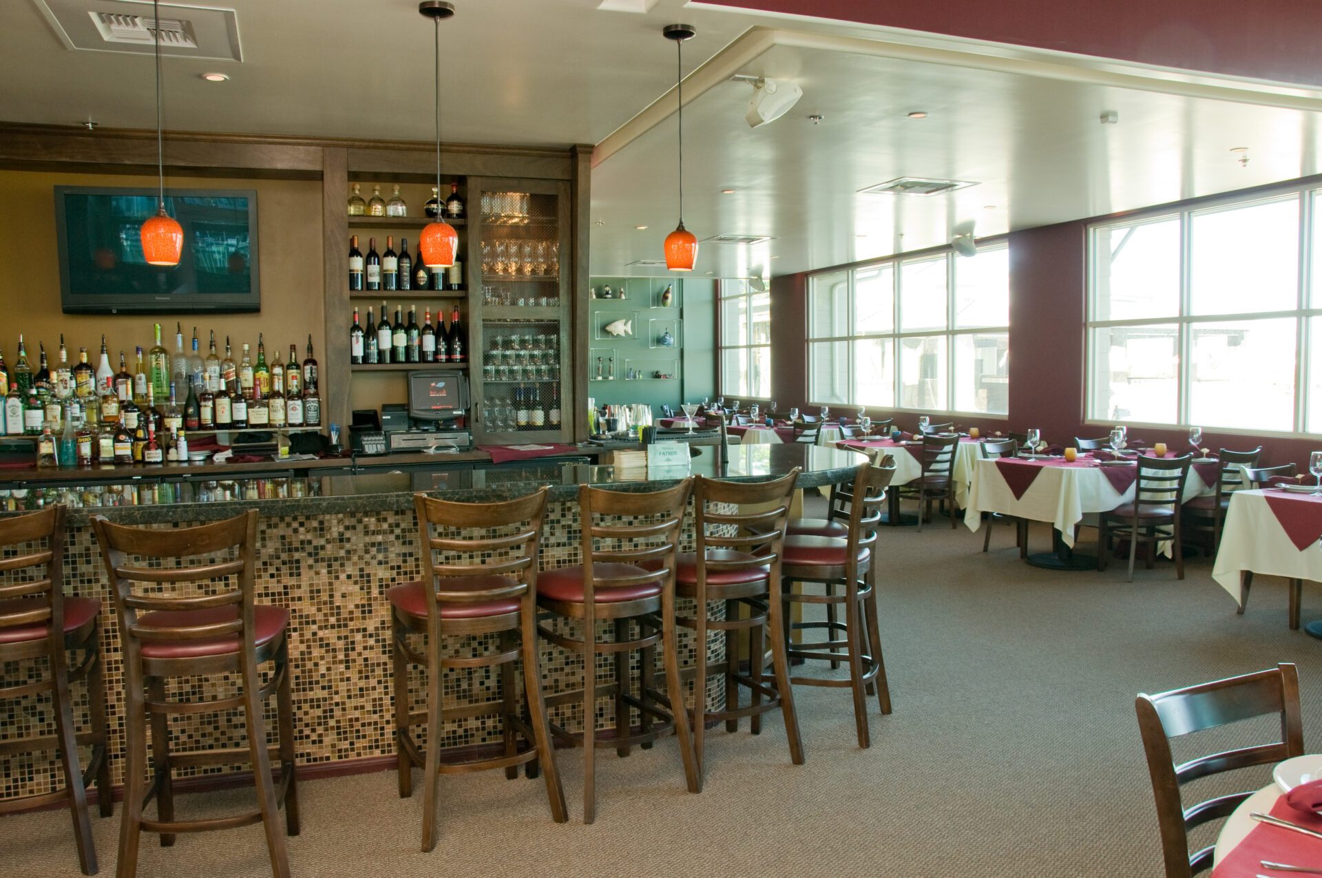 Restaurant interior with a bar counter lined with wooden barstools. Behind the bar are shelves with various bottles and glasses. The dining area features tables covered with white and burgundy tablecloths, set with glasses and plates. Large windows provide natural light.