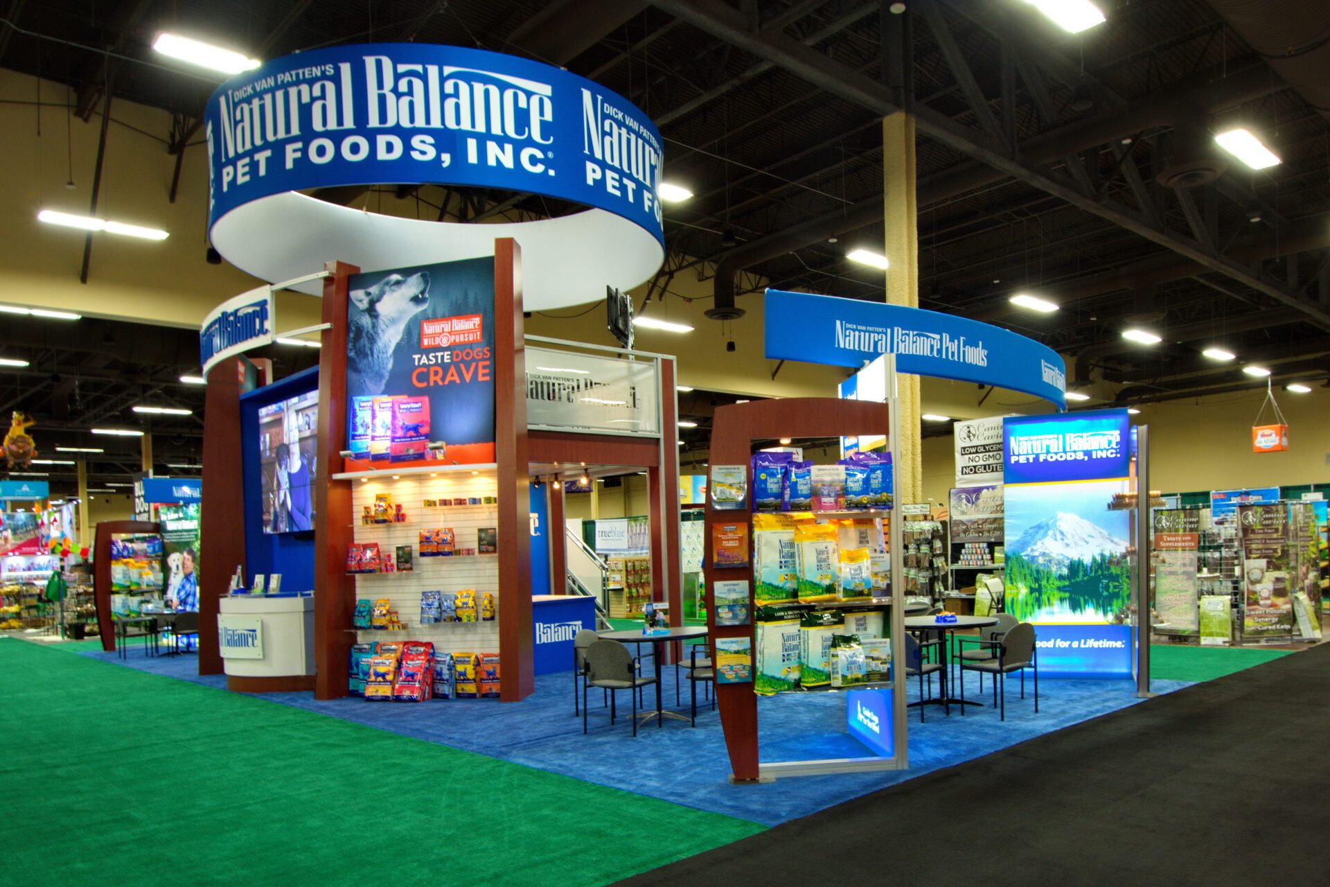 A trade show booth features Natural Balance Pet Foods, Inc. branding. The setup includes promotional materials, product displays, and seating areas. Large blue and white signs hang above, showcasing various pet food products. The background shows other exhibition booths.