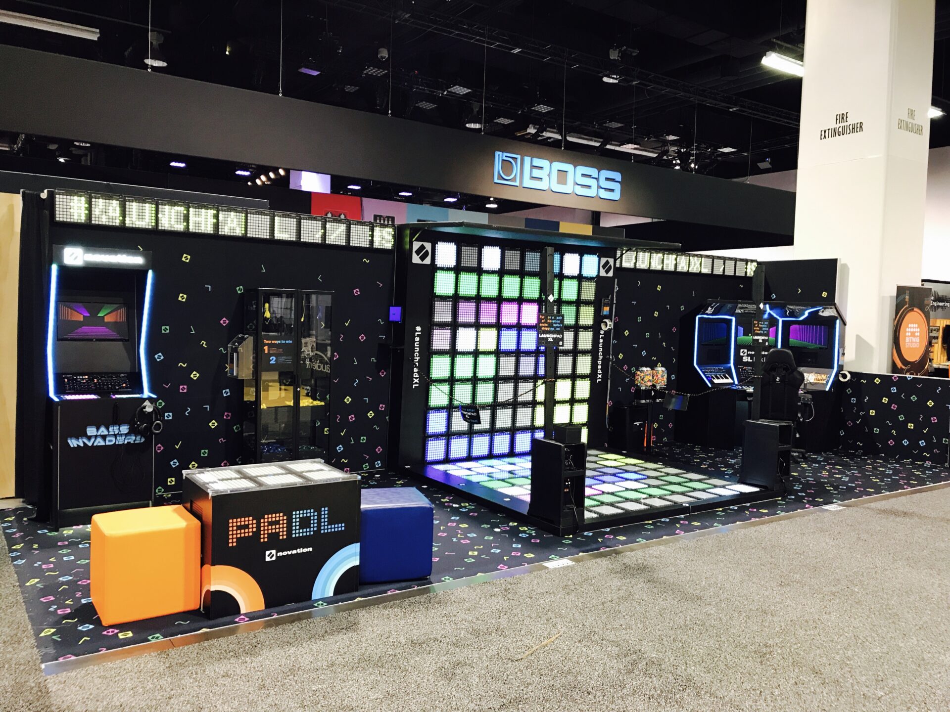 A gaming booth at a convention features multiple retro arcade game machines, including "Bass Invaders" and "Paddle." The setup includes a colorful LED-lit dance floor and seating cubes. The booth is branded with "BOSS" signage above.