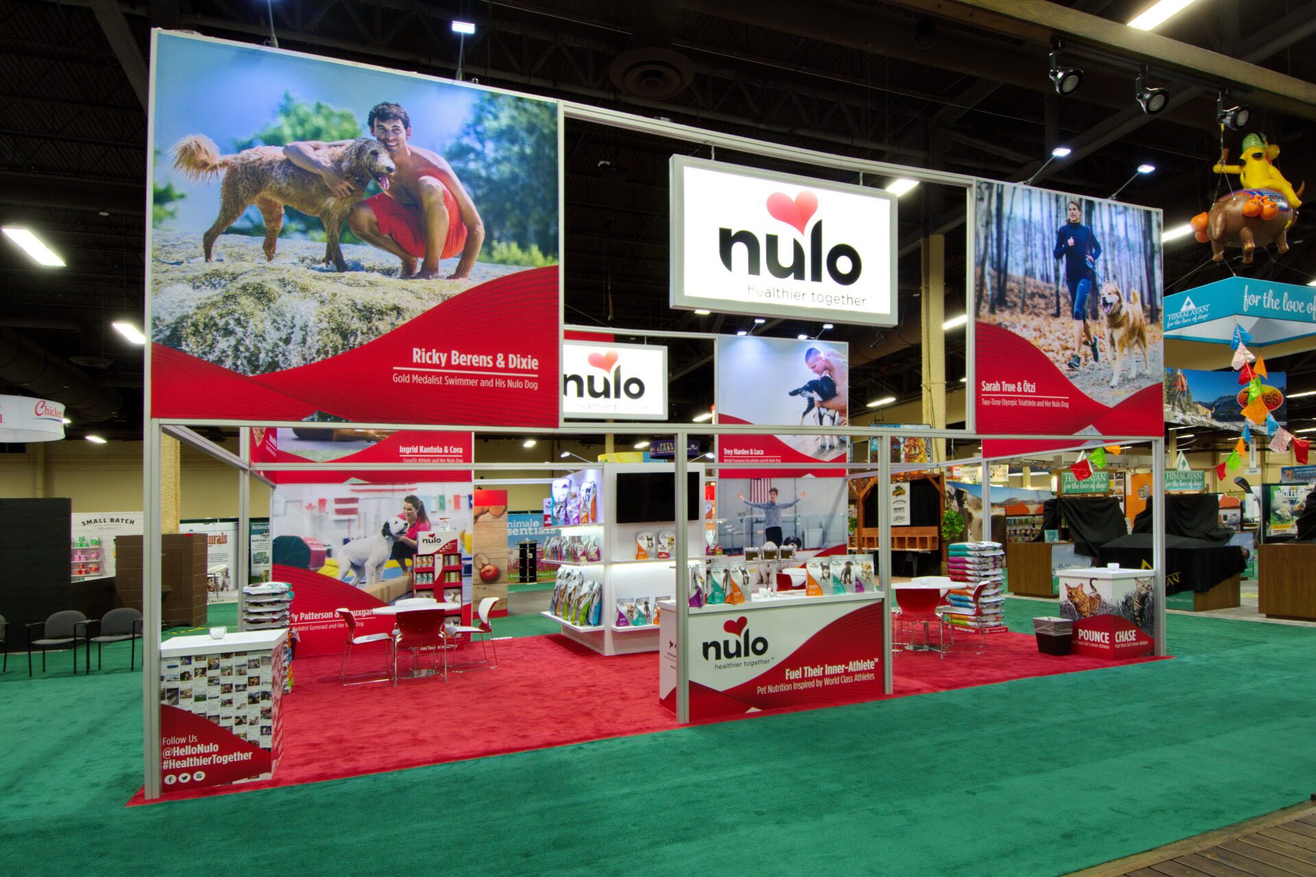 A trade show booth with "Nulo" branding. There are banners with images of dogs and people exercising. The setup includes tables, chairs, and product shelves. Prominent colors are red, white, and green. A sign reads "Feed Their Inner Athlete," promoting canine nutrition.