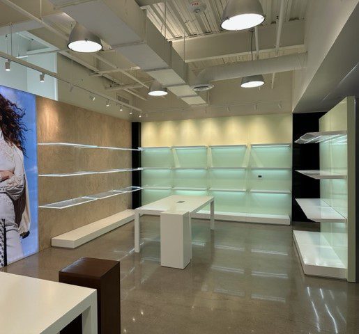 A modern retail store interior features well-lit, empty shelves and display areas on polished concrete floors. There is a large poster on one wall, a central display table, and bright overhead lighting, creating an airy and clean atmosphere.