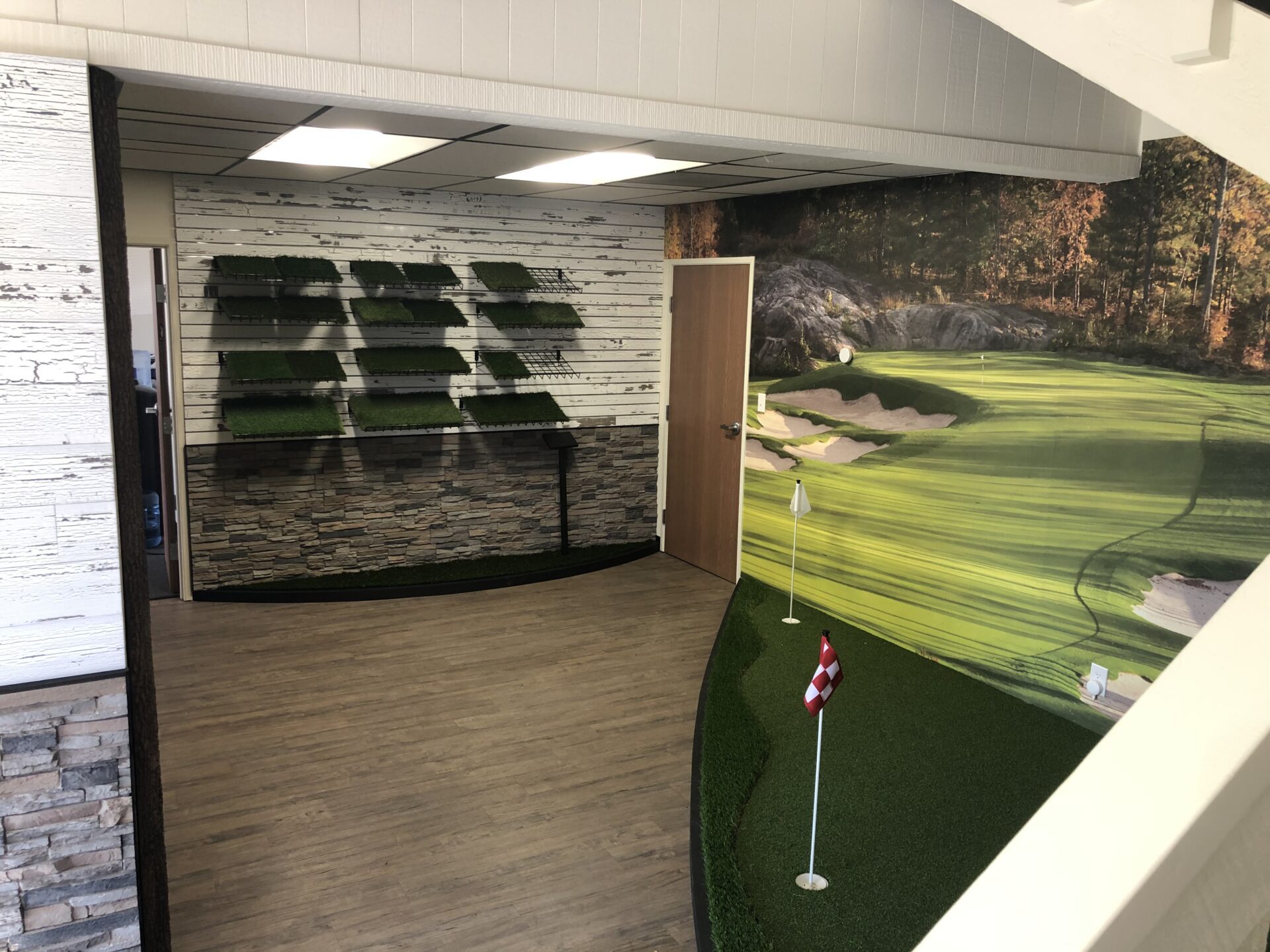 A minimalist golf store interior with a small putting green setup, various artificial grass samples on shelves, and a scenic golf course wall mural. Light wood flooring and stone accents create a modern and inviting atmosphere.