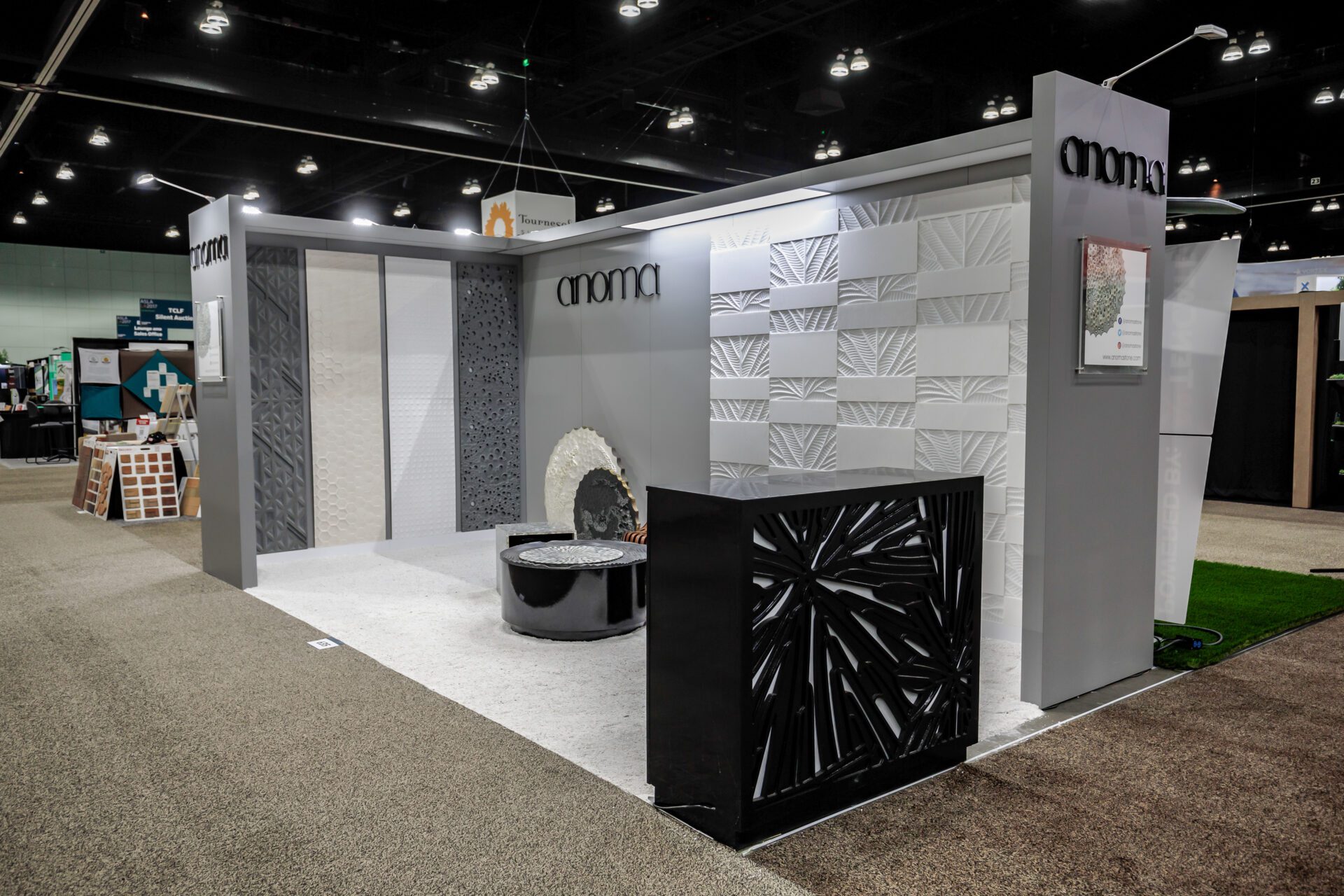 A modern trade show booth with the name "Aroma" displayed prominently features decorative wall panels with intricate patterns. A round seating area with a black and white mosaic design is in front, accompanied by a black counter with a geometric design.