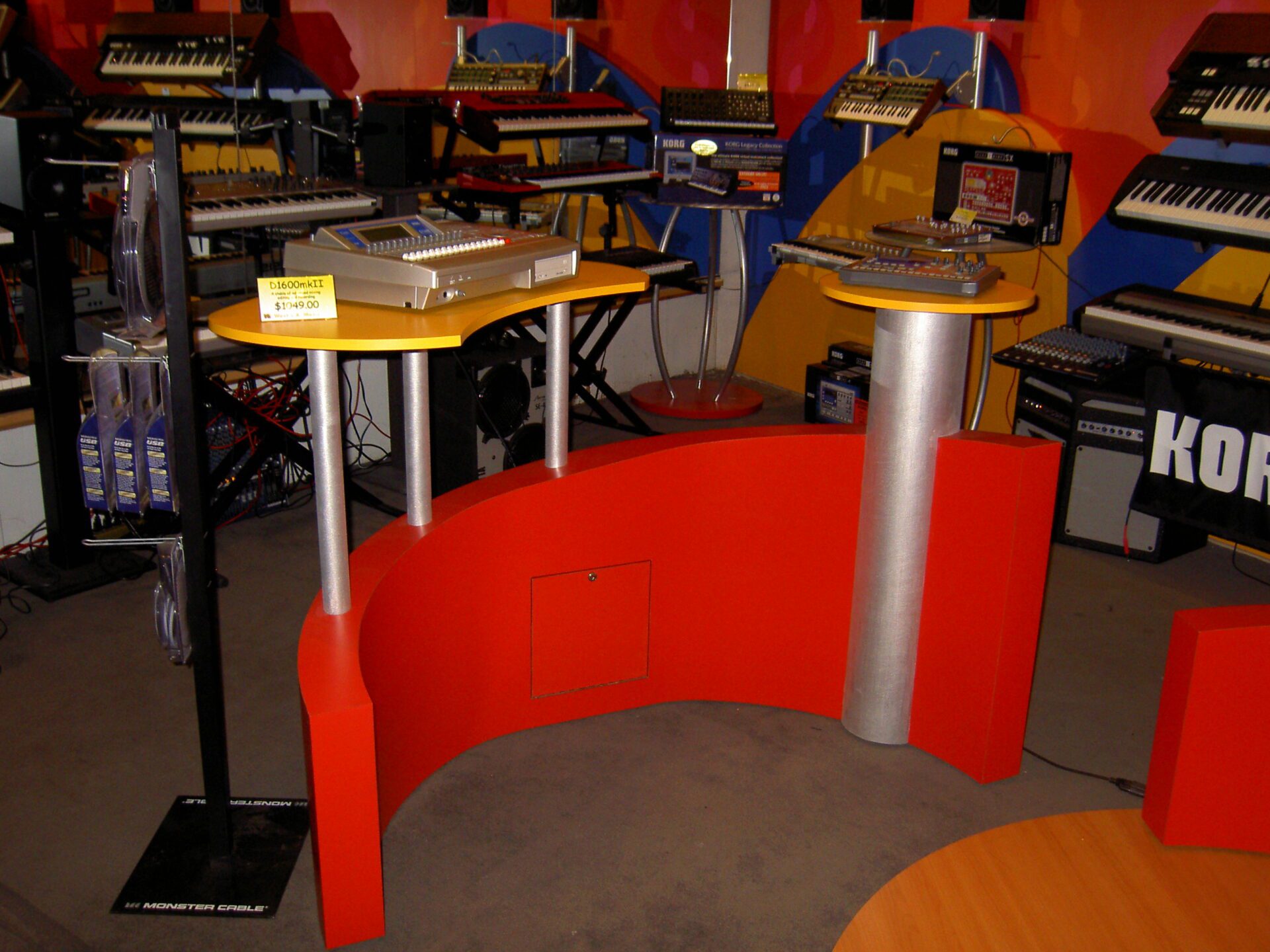 A brightly lit music store display featuring multiple electronic keyboard synthesizers on colorful stands. In the foreground, a vibrant red and yellow curved table showcases two sound mixing consoles. Background shelves are filled with various musical equipment.