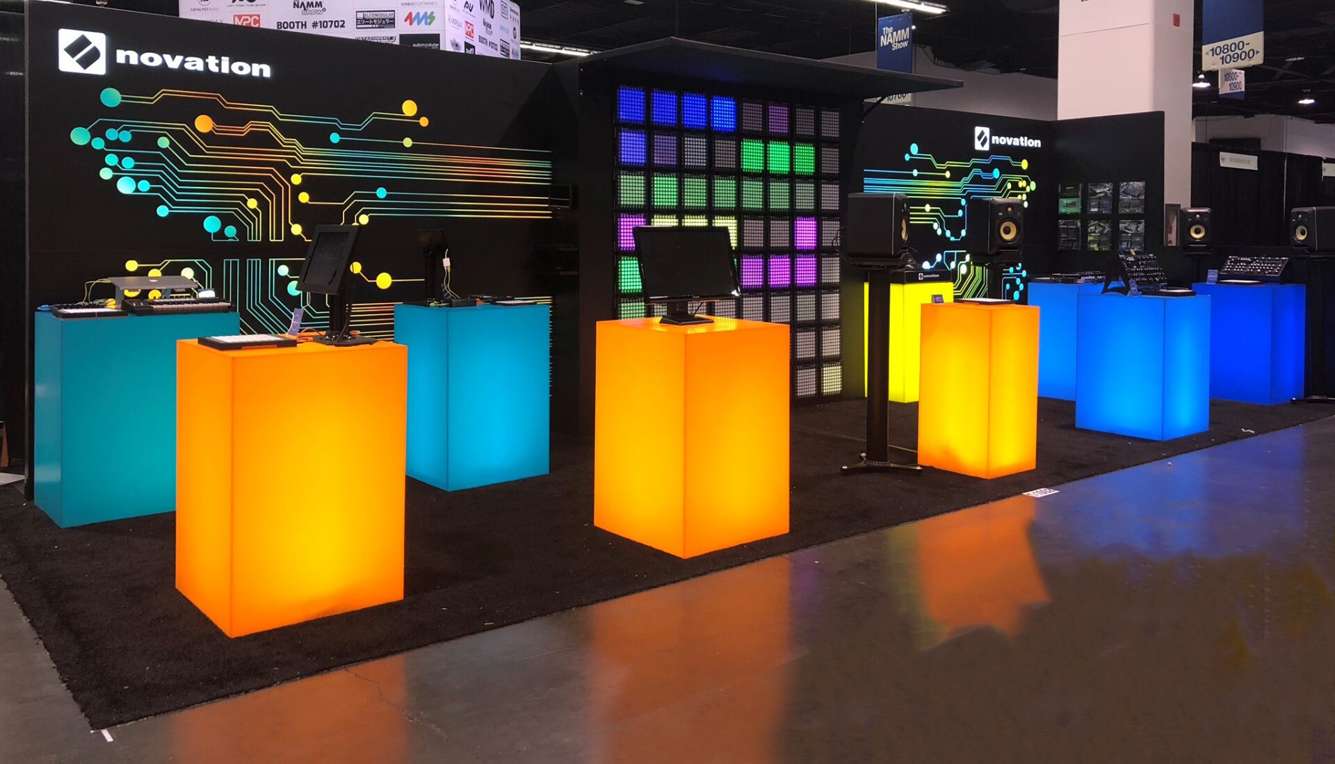 A trade show booth displays computer monitors on illuminated podiums in vibrant colors: orange, light blue, yellow, green, and blue. The back wall features colorful circuit-like designs and a digital screen grid, with "Novation" branding visible.