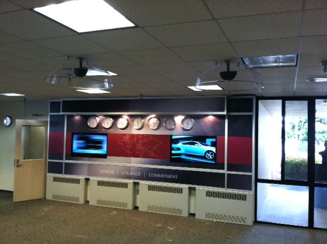 A wall display with several clocks showing different time zones, a map of the world in the center, and two screens below showing a car and other content. The wall has the words "Honor," "Courage," and "Commitment." The room has fluorescent lighting and a glass door.