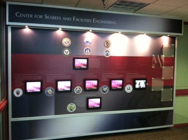 A display wall titled "Center for Seabees and Facilities Engineering" with multiple screens and insignias of various military branches. The wall is decorated in red, white, and blue and provides informational text and multimedia content about Seabees and engineering.