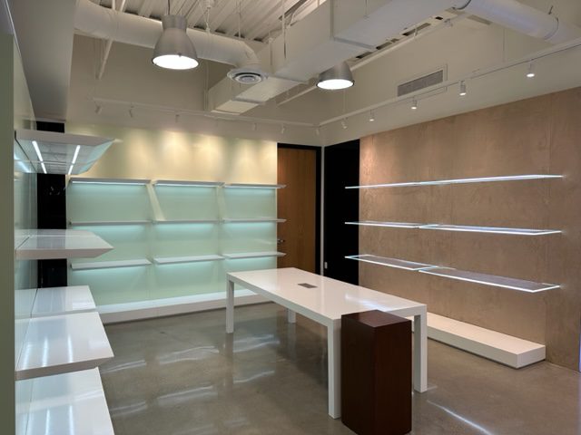 A modern, empty retail store with white shelves on light green and beige walls. The space features a large white table in the center with a small brown counter next to it. Exposed industrial-style ceiling and polished concrete floors complete the minimalist design.