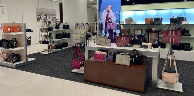 A neatly arranged display of designer handbags in various styles and colors is showcased in a well-lit store. Shelves and tables feature purses and totes, with a life-sized promotional image of a person carrying one of the bags in the background.