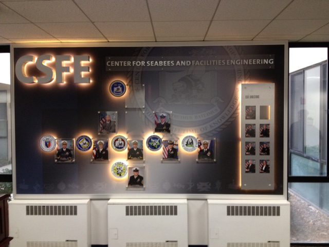 A wall display in an office features the illuminated letters "CSFE" with the words "Center for Seabees and Facilities Engineering" beneath. The display includes various Seabees insignias, text, and photos of individuals arranged in a grid pattern.